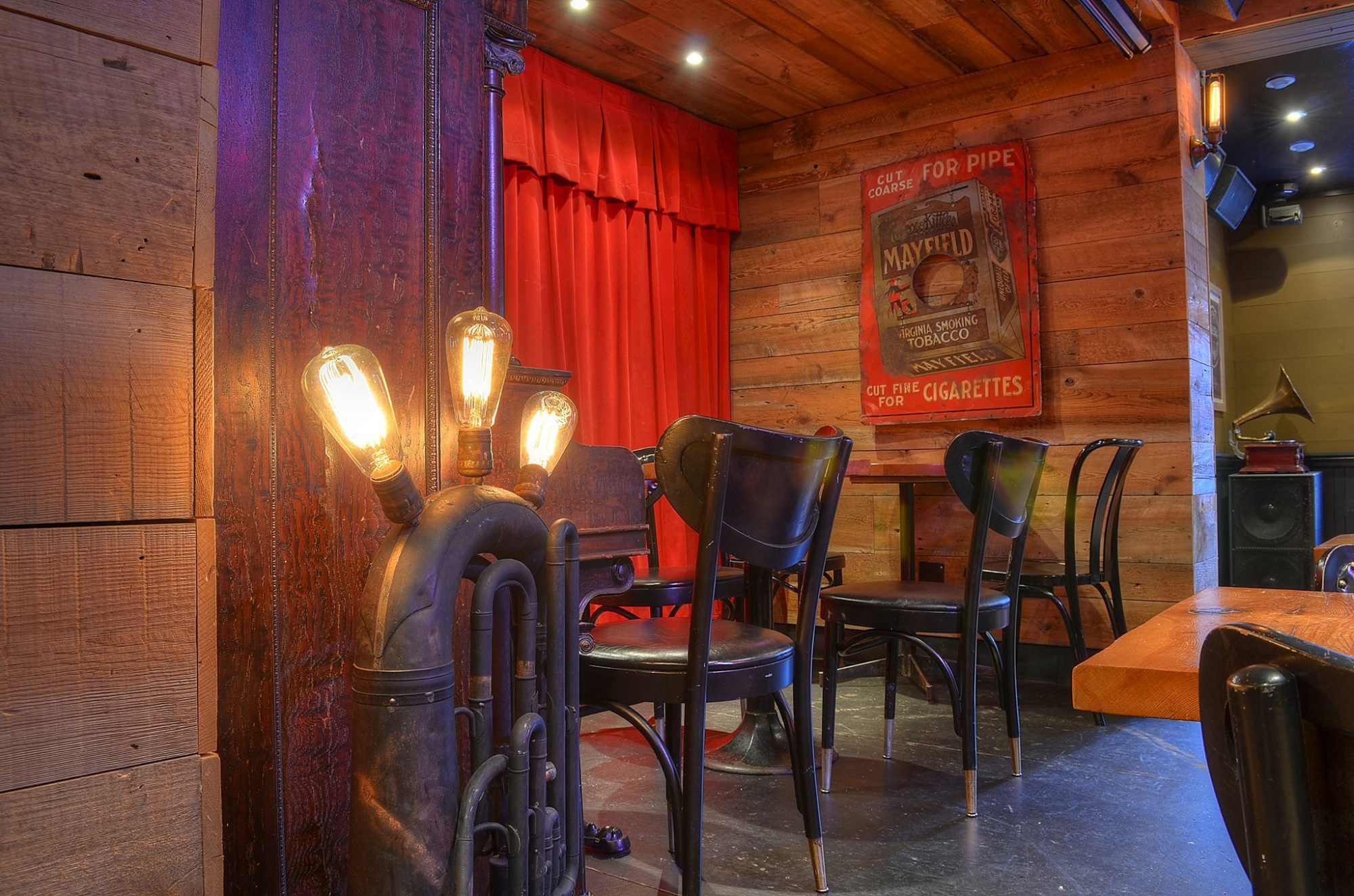 L'image montre un coin d'un bar ou d'un restaurant au style rétro. Le décor est composé de bois vieilli, de chaises noires aux pieds dorés, d'une publicité vintage pour du tabac Mayfield, d'un gramophone et d'un luminaire original fait à partir d'un tuba. Une partie du décor est cachée par un rideau rouge. L'ambiance est feutrée et évoque les années 1920-1930. Le contraste entre le bois brut et les éléments métalliques crée une atmosphère chaleureuse et stylisée. L'éclairage tamisé ajoute au charme rétro de la scène.