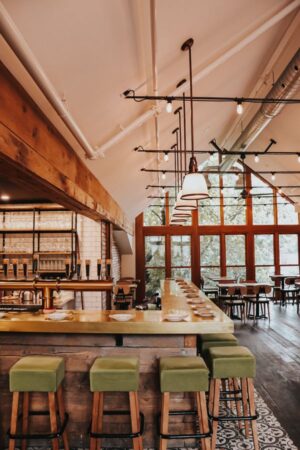 L'image montre l'intérieur d'un bar ou d'un restaurant élégant. Le comptoir, en métal doré, est long et doté de tabourets hauts aux sièges vert olive. Derrière le comptoir, on voit un système de tireuse à bière. L'espace est lumineux grâce à une grande baie vitrée donnant sur une végétation extérieure. Le style est industriel-chic, avec des poutres en bois apparentes, des tuyaux métalliques et des luminaires suspendus. Le sol est en bois, avec des carreaux décoratifs près du comptoir. Des tables et des chaises sont disposées dans la salle à manger, créant une ambiance chaleureuse et accueillante.