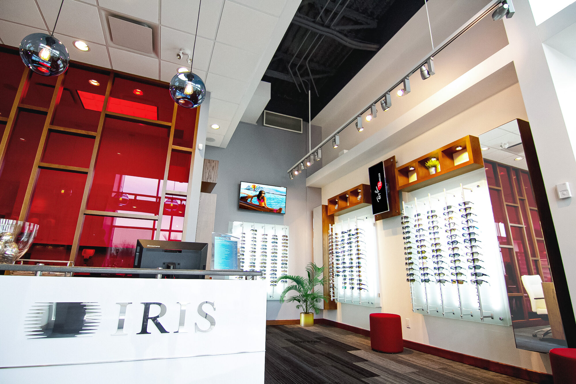 Aménagement moderne d'une clinique optométrique : comptoir d'accueil blanc avec dessus en granit noir, mur rouge lumineux avec structure en bois, éclairage suspendu design et présentoirs de lunettes rétroéclairés. L'ensemble crée une ambiance professionnelle et accueillante, alliant fonctionnalité et esthétique contemporaine.
