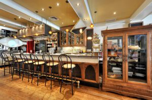 L'intérieur du bar de la Brasserie Chez Lévêque est élégant, avec des chaises à haut dossier au comptoir, des lampes suspendues, des étagères avec des bouteilles, de la verrerie au-dessus de la tête, et une vitrine en bois remplie de pâtisseries et d'assiettes sur un sol en bois poli.