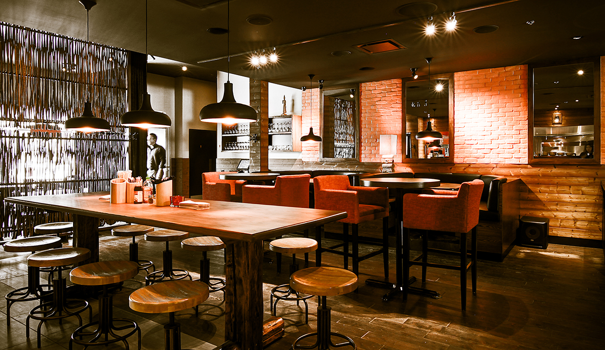Un intérieur de restaurant moderne avec des tables en bois, des tabourets et des chaises rouges rembourrées. Un éclairage chaleureux est suspendu au plafond et une personne se tient près de l'espace bar. Une ambiance classique que l'on retrouve dans les Restaurants Sésame, avec des murs en briques et en bois.