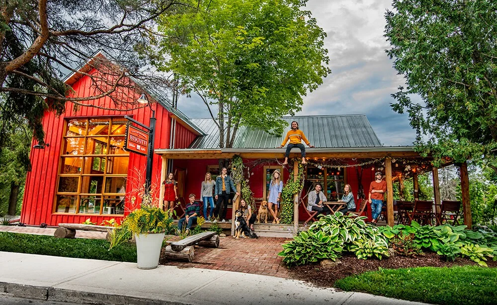 Un groupe de personnes et un chien se rassemblent à l'extérieur du bâtiment rouge vif de Biscotti & Cie Old Chelsea, avec de grandes fenêtres et un porche, entouré d'arbres et de plantes vertes sous un ciel nuageux. Une personne est assise sur le toit au-dessus du porche.