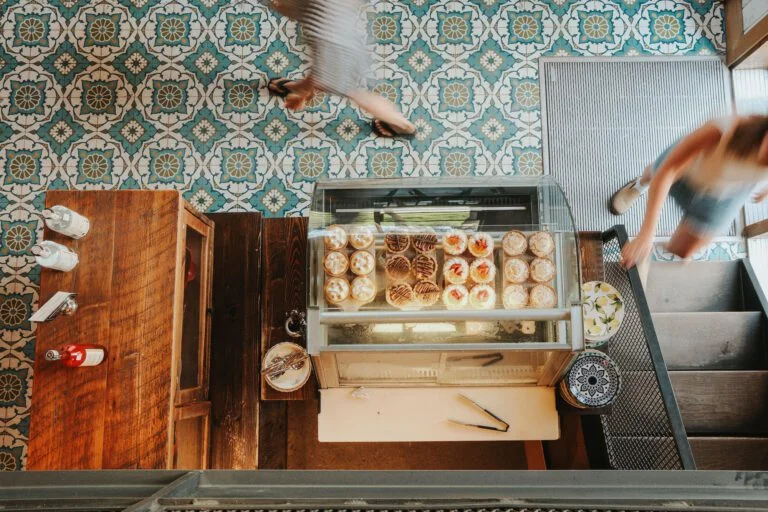 Vue aérienne de la vitrine de la boulangerie Biscotti & Cie Old Chelsea avec des pâtisseries assorties, un comptoir en bois avec des condiments, et deux personnes floues marchant sur un sol carrelé décoratif.