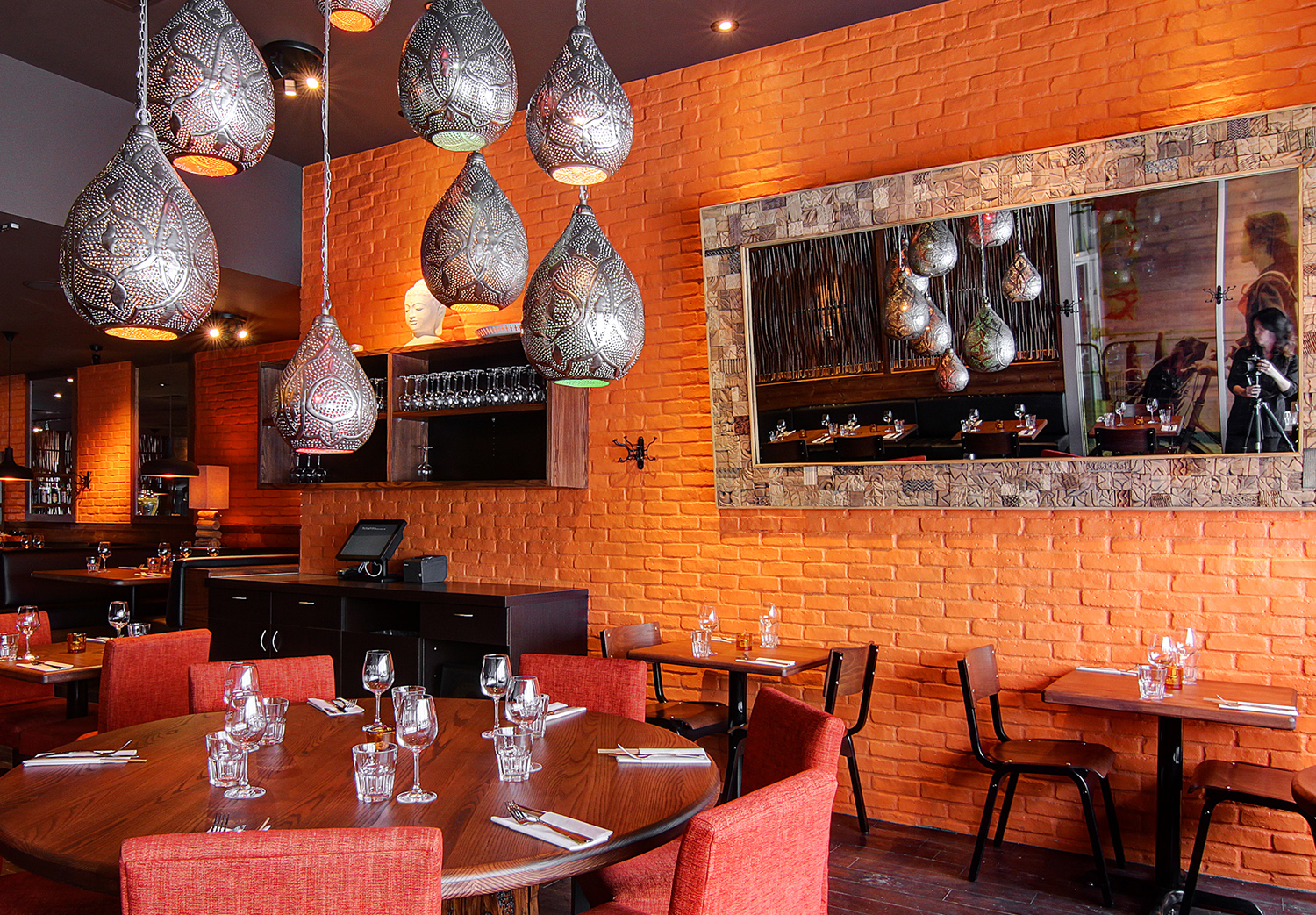 Le restaurant chaleureux est doté dun aménagement bien pensé, avec des murs en briques orange chaleureux. Des suspensions lumineuses projettent une douce lueur sur les tables dressées avec des verres et des couverts. Un grand miroir décoratif renforce la profondeur, tandis que les chaises rouges complètent à merveille les tons terreux.