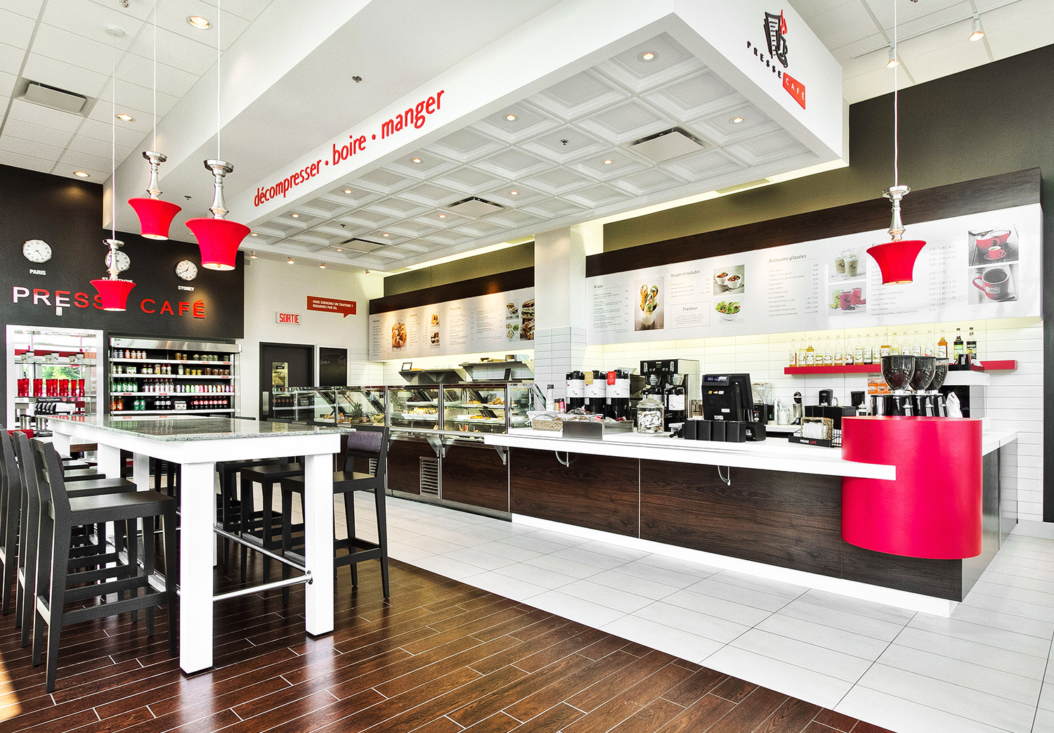 Intérieur lumineux d'un café-restaurant Presse Café, au design commercial moderne. Grand comptoir de service en bois et blanc avec menu rétroéclairé, présentoirs vitrés pour viennoiseries et sandwiches, et espace café avec machines à espresso. Mobilier épuré avec table haute et chaises noires, éclairage suspendu avec luminaires rouges emblématiques. Sol en bois et carrelage blanc contrastant, créant une ambiance dynamique et accueillante.