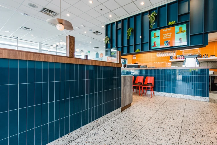 Modern fast casual restaurant interior with blue tiled counter, terrazzo floor, orange chairs, menu screens, and plants on the wall.