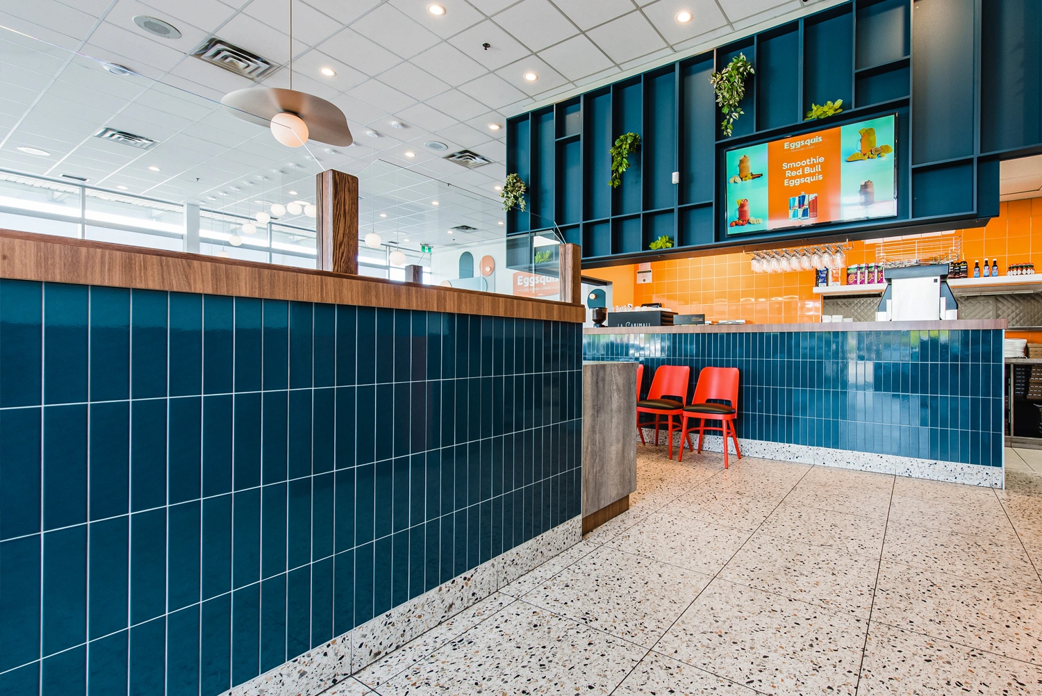 Modern fast casual restaurant interior with blue tiled counter, terrazzo floor, orange chairs, menu screens, and plants on the wall.