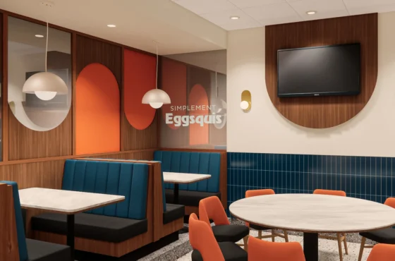 Salle intérieure d’un restaurant moderne avec des banquettes bleues et des panneaux muraux en bois ornés de formes orange et blanches. Des luminaires arrondis suspendus éclairent les tables en marbre clair. À droite, des chaises orange entourent des tables en bois et une télévision est fixée au mur. L’ensemble crée une ambiance chaleureuse signée Dany Perron.