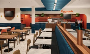 Vue intérieure d’un restaurant moderne aux couleurs vives, avec des banquettes bleues, des tables en marbre clair et des chaises en bois. Le comptoir de service au fond est décoré de carreaux orange, de plantes vertes et d’étagères en bois. Deux personnes se tiennent au comptoir tandis qu’un éclairage rond suspendu illumine l’espace. L’ambiance est chaleureuse et organisée dans un style Mid Century revisité.