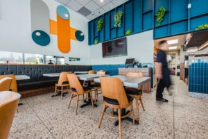 Vue sur un restaurant au décor minimaliste et coloré, avec des assises confortables, des tables carrées et un agencement mural bleu rehaussé de plantes vertes.