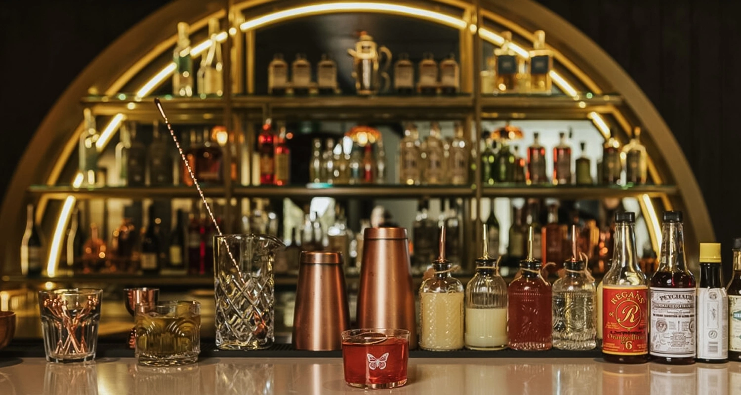 Un comptoir de bar élégant avec des shakers, des verres à mélange, des bitters et des bouteilles, placés sur des étagères éclairées remplies de liqueurs et de verrerie, créant une atmosphère haut de gamme et invitante.