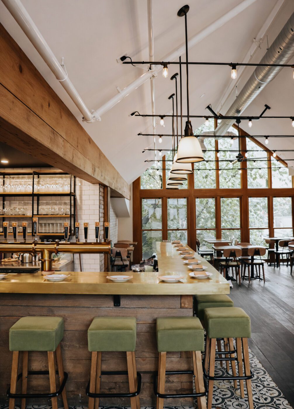 Un bar moderne avec un long comptoir, des tabourets verts, des lampes suspendues, de grandes fenêtres laissant entrer la lumière naturelle et des éléments en bois. Des assiettes et des verres sont posés sur le comptoir et des tables sont disposées à l'arrière-plan.
