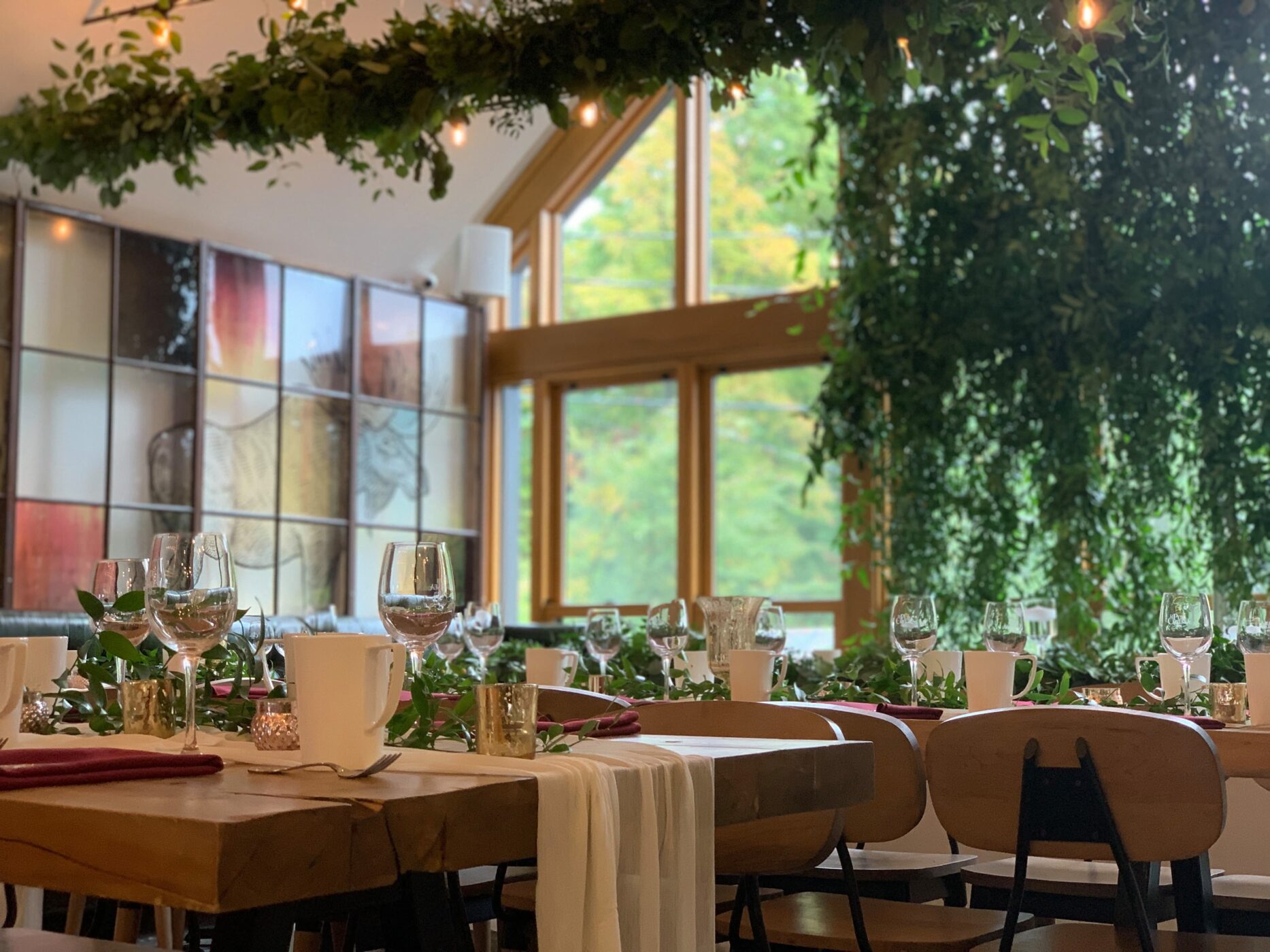 Une table à manger joliment dressée dans une pièce lumineuse avec de grandes fenêtres, de la verdure suspendue au plafond, des verres, des assiettes et des serviettes soigneusement disposés, créant une atmosphère élégante et festive.