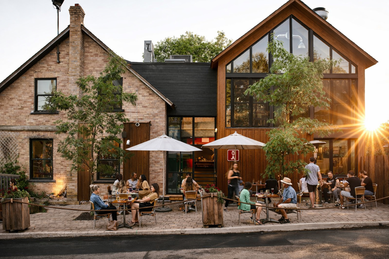 Des personnes sont assises à des tables en plein air sous des parasols devant un café rustique moderne avec de grandes fenêtres et des accents en bois, entouré d'arbres et de plantes, alors que le soleil se couche en arrière-plan.